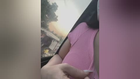 Media: Video of a person in a pink ribbed top and black dress, sitting in a car looking out the window at a sunset scene.