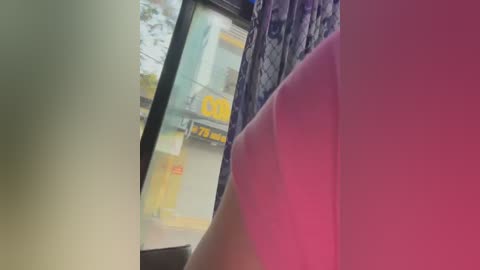 Media: A blurry video of a window with a yellow sign reading \"7-11\" and a partially visible person in a pink shirt. The background includes a colorful curtain and a glimpse of a building outside.