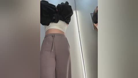 Media: Video of a person wearing a white crop top and loose, light gray sweatpants, standing in a narrow, bright hallway. Their arms are raised, revealing a black jacket tied around their waist.