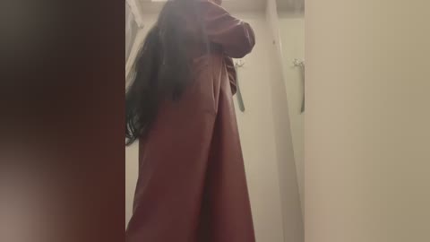 Media: A video of a person with long, dark hair, wearing a burgundy robe, standing in a dimly lit, beige-walled bathroom. The image is slightly out of focus, emphasizing a candid, private moment.