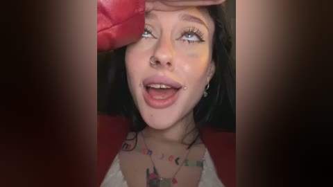 Media: Video of a young woman with pale skin, blue eyes, and black hair, wearing a red top, a nose ring, and a tongue piercing, looking up with a playful expression.