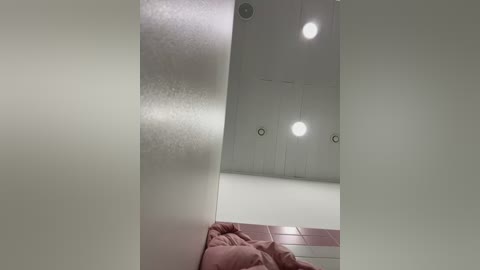 Media: Video of a minimalist, modern room with white walls and ceiling. Three round, white pendant lights hang from the ceiling. A large, rectangular mirror divides the scene, reflecting the room's interior. A pink blanket is visible in the foreground, adding a soft contrast to the stark white decor.