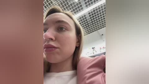 Video of a young woman with light skin, straight blonde hair, and a septum piercing, making a pouty face, taken in a dimly lit room with a tiled ceiling and colorful, abstract artwork on the wall.