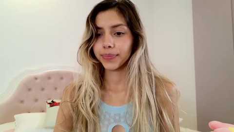 Media: Video of a young Latina woman with long, wavy, light brown hair, wearing a light blue lace bralette, sitting on a bed with a pink, tufted headboard.