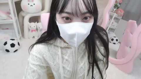 Media: Video of an Asian woman with long black hair, wearing a white face mask and a cream knitted sweater, in a pastel-colored, bunny-themed room with plush toys and a pink gaming chair.