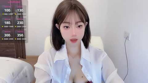 Media: Video of an East Asian woman with shoulder-length brown hair, wearing a white blouse unbuttoned to reveal cleavage, sitting on a bed in a minimalist room.