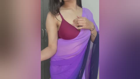 Media: Video of a South Asian woman in a purple saree, revealing a maroon bra. She has medium skin tone, dark hair, and a tattoo on her left arm. Background features a gray door and white wall.