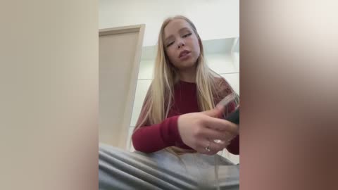 Media: Video of a young woman with long blonde hair, wearing a red sweater, holding a phone in a dimly lit room with beige walls and a door.