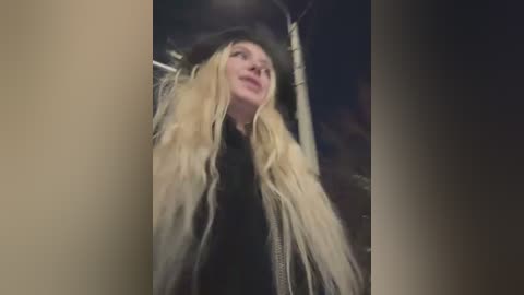 A video of a blonde woman with long hair, wearing a black hat and jacket, looking upward in a dimly lit, nighttime cityscape.