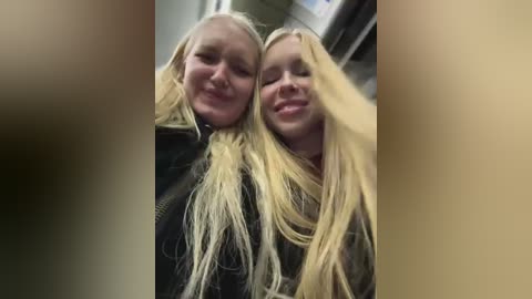 Video of two smiling blonde women with long hair, wearing black jackets, standing close together in a dimly lit room, with blurred background.