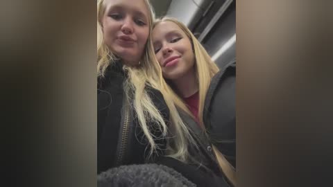 Media: Video of two young blonde women with straight hair, smiling closely together. One wears a black jacket, the other a red shirt. They're indoors, possibly on a train, with blurred background.