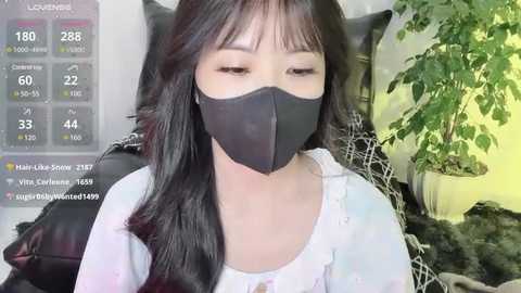 Media: A video of an East Asian woman with long black hair, wearing a black face mask and a white blouse, seated indoors, with a green plant and digital weather display in the background.