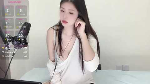 Media: Video of an Asian woman with long black hair, wearing a white dress, holding a microphone, in a simple bedroom.