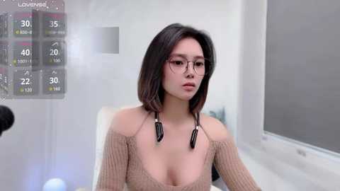 Media: Video of a young East Asian woman with straight black hair, wearing glasses, a low-cut beige knit top, and black headphones, sitting indoors.