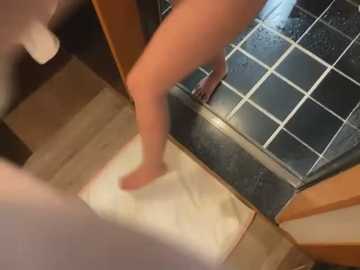 Media: A video captures a nude person's legs stepping into a shower with a black and white tiled floor. The individual has light skin, and the setting is a bathroom with wooden accents.