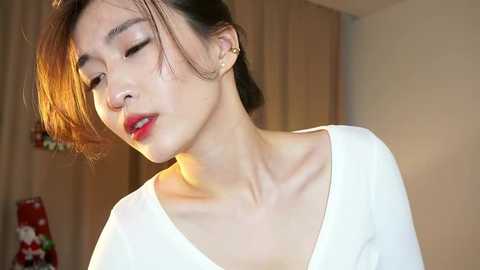 Video of an Asian woman with short, light brown hair, wearing a white top, with closed eyes and slightly parted lips. She has a slender build and fair skin. Background includes a beige curtain and a decorative doll.