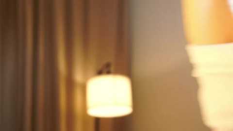 A blurred video of a warm-toned, cylindrical pendant light hanging from a beige wall. The background features a soft, neutral-colored curtain. The image has a cozy, ambient lighting effect.