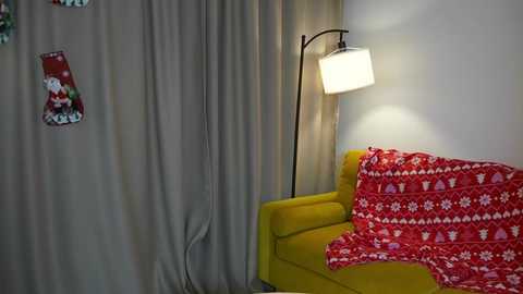 Video of a cozy living room corner with a mustard-yellow armchair draped in a red and white patterned blanket, a modern black floor lamp, and beige curtains.