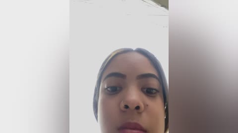 Media: Video of a young woman with medium-brown skin, dark brown eyes, and light brown hair, wearing a nose piercing, taken from a low angle, focusing on her face.