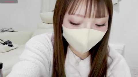 Media: Video of a young Asian woman with long brown hair, wearing a white face mask and robe, sitting in a spa chair, surrounded by white decor.