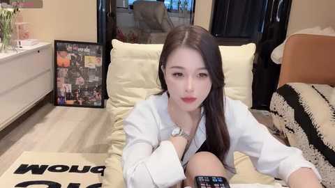 Media: A video of an East Asian woman with long black hair, wearing a white shirt, sitting on a beige cushion, surrounded by a modern living room with a framed collage, a TV remote, and a patterned blanket.