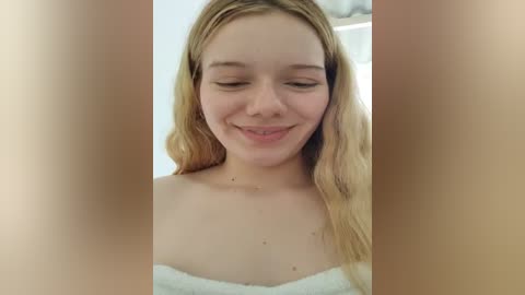 Media: A video of a young Caucasian woman with fair skin and long, wavy blonde hair, smiling softly. She wears a white towel wrapped around her shoulders, suggesting she is fresh out of the shower. The background is blurred, focusing attention on her.