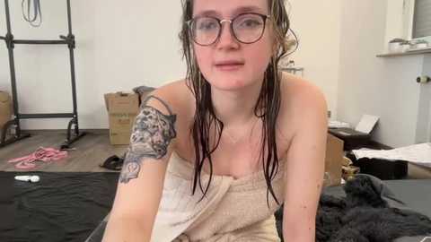 Media: Video of a fair-skinned, wet-haired woman with glasses and a large tattoo on her upper arm, wrapped in a towel, sitting on a black table in a messy, industrial-style room.