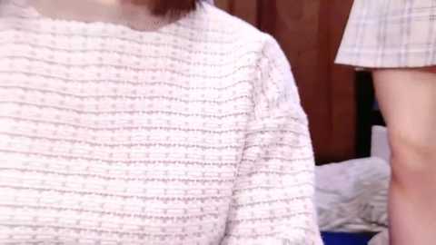 Media: Video of a person with shoulder-length brown hair wearing a white, textured, long-sleeved shirt with thin, horizontal stripes, standing in a room with a wooden background and a bed with white sheets.