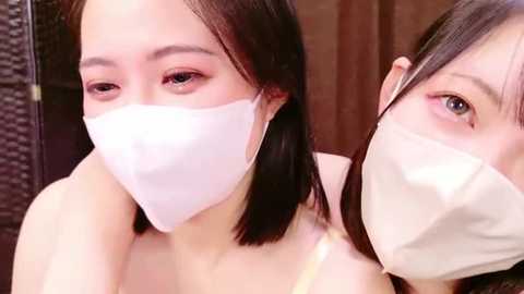 Media: Video of two young East Asian women with pale skin, wearing white medical masks, brown hair, and pink eyeshadow, standing close together, indoors.