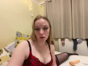 Media: Video of a fair-skinned, blonde woman with a neutral expression, wearing red lace lingerie and black stockings, sitting on a bed in a dimly lit room with yellow walls, beige curtains, and string lights.