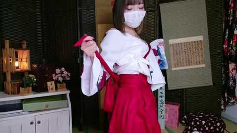 Media: Video of an Asian woman in a traditional Japanese kimono with red and white colors, white mask, and red obi, holding a red umbrella, standing in a dimly lit room with wicker furniture and Japanese decor.