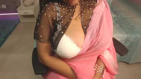 Media: Video of a woman in a sheer black blouse adorned with white polka dots, revealing a white lace bra underneath. She wears a pink sari, sitting on a modern chair in a room with a teal carpet and a white nightstand.