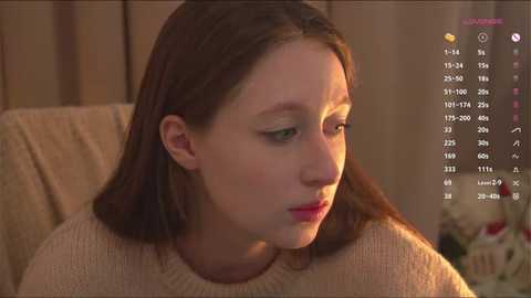 Video of a young Caucasian woman with fair skin, brown hair, wearing a beige sweater, looking contemplative, set in a dimly lit room with a calendar on the right.