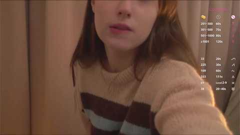 Video of a young woman with fair skin and brown hair, wearing a beige and brown striped sweater, partially visible face, dimly lit indoor setting, background showing a calendar interface.