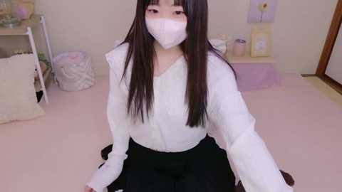 Media: Video of an East Asian woman with long black hair, wearing a white mask, white blouse, and black skirt, kneeling on a pink carpet in a softly lit, minimalist room with pastel decor.