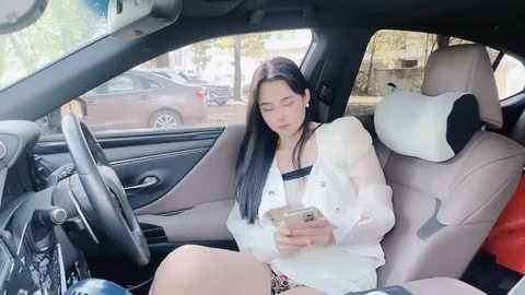 Media: Video of an Asian woman with long black hair, wearing a white blouse and shorts, sitting in a car, holding a phone, with a parked car visible outside the window.