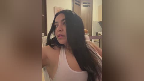 Media: Video of a young woman with long black hair, wearing a white tank top, standing in a dimly lit room with wooden furniture and a bed.