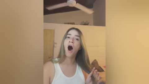 Media: Video of a young woman with light skin, long blonde hair, and a white tank top, mouth open in an exaggerated expression, standing in a dimly lit room with beige walls and a ceiling fan.