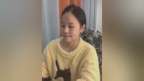 Media: Video of a young Asian girl with short black hair, wearing a yellow, fluffy pajama top with a black cat design, sitting indoors next to a patterned chair.