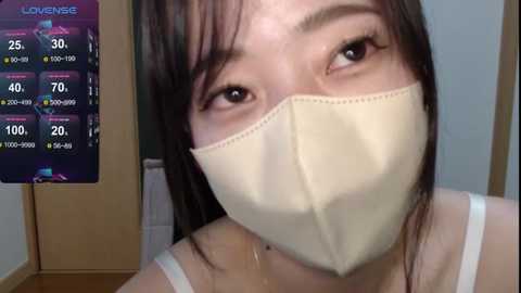 Media: A video of an Asian woman with light skin, wearing a beige medical mask, black hair, and a white tank top. Background includes a weather forecast screen and a wooden floor.