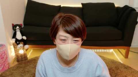 Media: Video of a short-haired Asian woman with pale skin, wearing a beige face mask, light blue sweater, sitting on a round brown rug in a modern living room with a black sofa, a stuffed cat, and a glowing lamp.