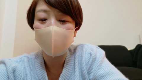 Media: Video of an Asian woman with short, dark brown hair, wearing a beige face mask and a light blue, knitted cardigan, sitting indoors with a black couch in the background.