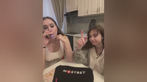 Media: Video of two young women in a kitchen. One with long brown hair, wearing a white tank top, brushes her teeth. The other, with short brown hair, in a beige hoodie, points with her right hand.