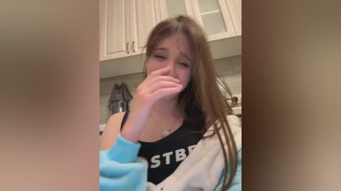 Media: Video of a young woman with long brown hair, wearing a black tank top with \"JUST BREATHE\" in white, sitting in a kitchen, covering her face with her hands, crying.