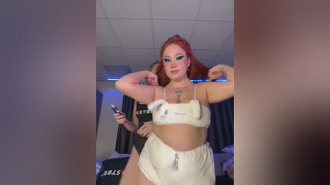 Media: Video of a young woman with red hair, wearing a white sports bra and shorts, flexing her bicep in a dimly lit room with a white ceiling and blue lighting.