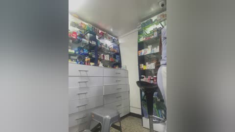 Video of a small, brightly lit pharmacy with white walls and a high shelf filled with colorful medicine bottles. A person in white scrubs stands at a black counter, while a metal stool is visible in the foreground.