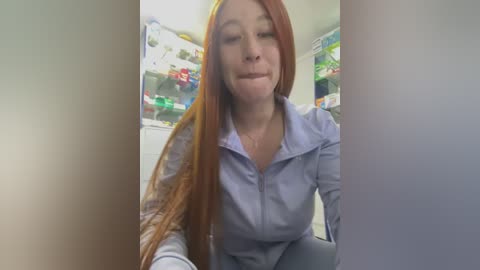 Video of a fair-skinned woman with long red hair, wearing a light blue button-up shirt, leaning against a fridge filled with colorful groceries.