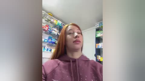 A video of a young woman with long red hair, wearing a purple hoodie, standing in a brightly lit convenience store with shelves full of colorful snacks.