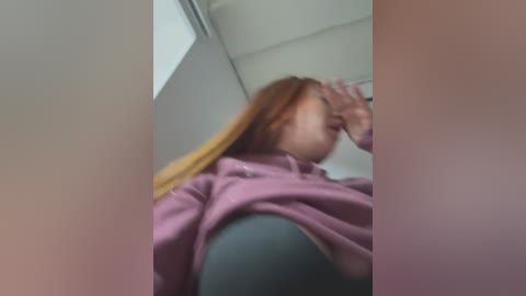 A blurred, candid video of a woman with long red hair, wearing a pink hoodie, covering her mouth with her hand, taken from a low angle in a bathroom with beige walls and a white ceiling.