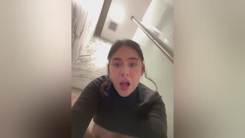 Media: Video of a young Asian woman with long black hair, wearing a black crop top, looking surprised, in a modern hallway with white walls, black-and-white patterned floor, and a metal railing.